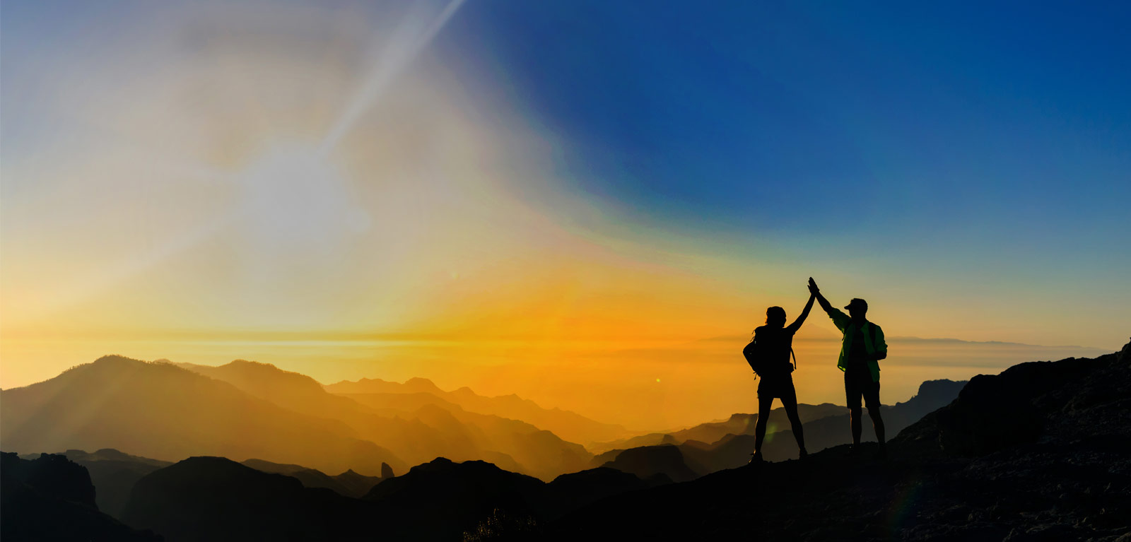 Two people, on top of a mountain high-five as they look into a sunset